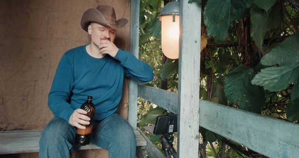 Cowboy man op veranda drinkt bier uit een fles en communiceert online via de telefoon - Foto, afbeelding