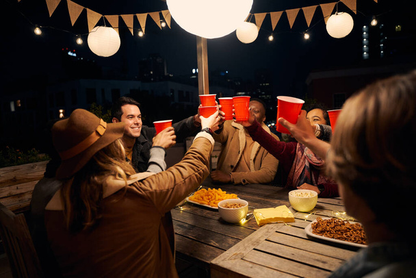 Group of diverse friends sitting, making a toast at an outdoor party around a boho themed table. Red cups and laughs - Fotó, kép