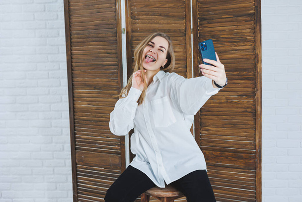 Emotional young woman in a white cotton shirt gesticulates and looks at the screen of a modern gadget. A girl with a mobile phone takes a selfie - Photo, Image