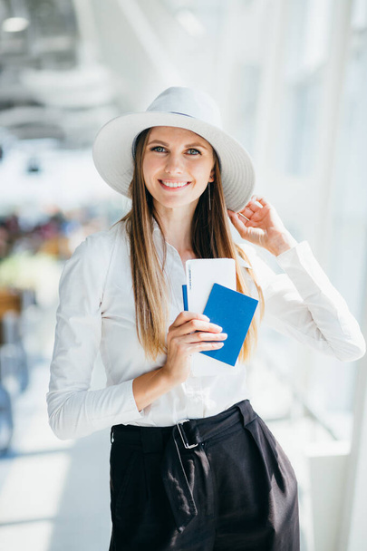 Веселая деловая женщина ждет свой рейс в аэропорту. Молодая женщина в аэропорту. Девушка с паспортом и посадочным талоном в аэропорту. Привлекательная молодая женщина отправляется в путешествие по воздуху - Фото, изображение