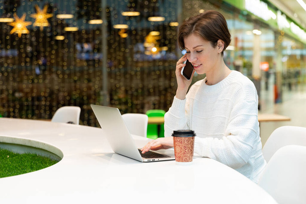 Una foto de una joven que se sienta en el patio de comidas de un centro comercial en una mesa y trabaja en una computadora portátil, habla por teléfono móvil. Concepto independiente - Foto, Imagen