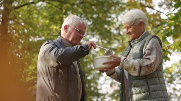 Un anciano europeo de cabello gris ayuda a los pobres sin hogar ofreciéndoles voluntariamente y vertiendo sopa nutritiva en el cuenco de plástico que pertenece a una anciana necesitada. - Imágenes, Vídeo