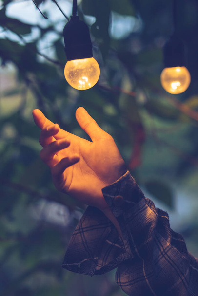 A close-up of the man's hand holding the light bulb replacing the old-fashioned orange light bulb The vintage hanging decoration in the park looks beautiful, the home design decorated with orange bulbs. - 写真・画像