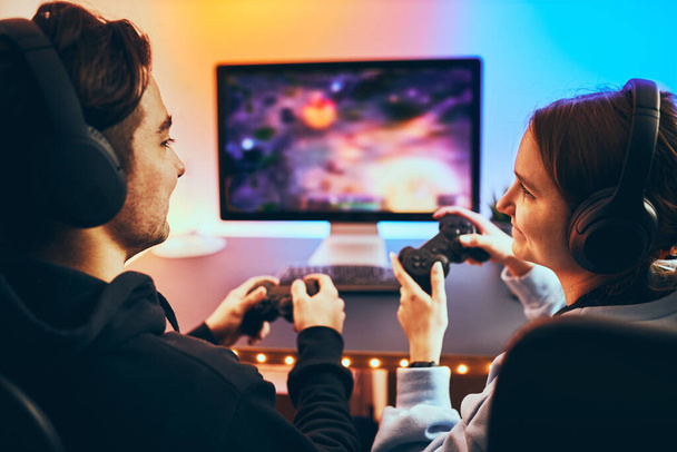 Friends playing video game at home. Gamers holding gamepads sitting at front of screen. Streamers girl and boy playing online in dark room lit by neon lights. Competition and having fun - Foto, Bild