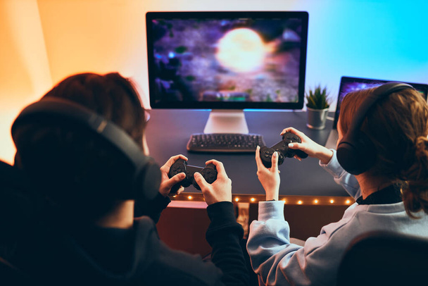 Friends playing video game at home. Gamers holding gamepads sitting at front of screen. Streamers girl and boy playing online in dark room lit by neon lights. Competition and having fun - 写真・画像