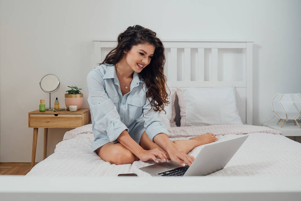 Fröhlich lächelnde Geschäftsfrau im Hemd sitzt auf einem Bett und tippt morgens etwas auf ihre Laptop-Tastatur - Foto, Bild