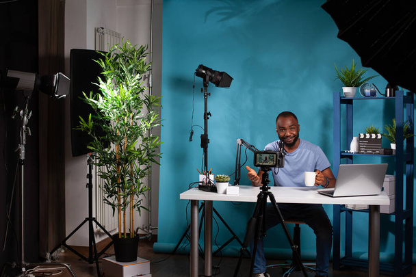 Создатель контента разговаривает с последователями перед съёмками видеокамеры на штативе, сидя за столом - Фото, изображение