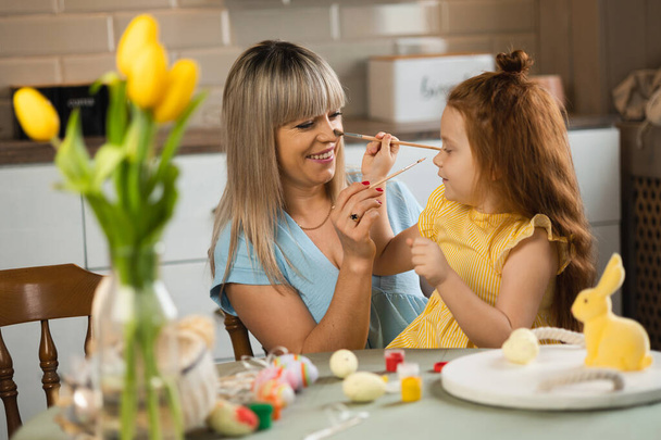 Mãe e filha se divertem brincando com pincéis na cozinha à mesa enquanto pintam ovos para a Páscoa - Foto, Imagem