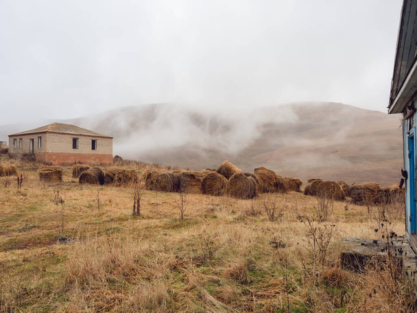 Área alpina com palheiros perto de casas abandonadas entre grama seca de outono e colinas nubladas ao redor - Foto, Imagem