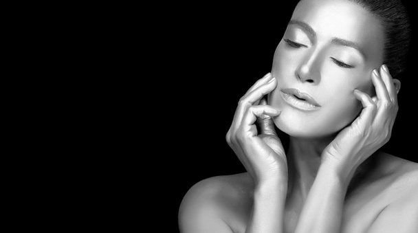 Silver skin model girl. Beautiful sensual young woman with glowing metallic silver body makeup raising hands to her face with eyes closed and a serene expression. Monochrome portrait isolated on black - Zdjęcie, obraz