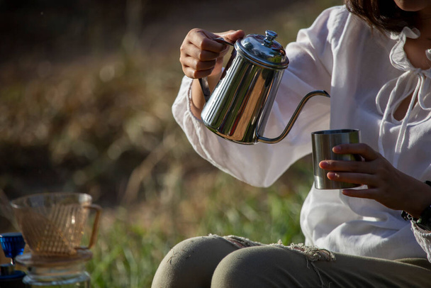 jarra de termo de acero inoxidable En las manos de una mujer asiática vertiendo café en una taza en una taza de acero inoxidable. con sol de la mañana Después de acampar y descansar en la mañana - Foto, Imagen
