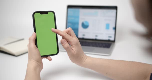 Chroma Key Mock-Up op Smartphone in hand.Vrouw met behulp van telefoon met groen scherm. Laptop met grafieken op het scherm en notitieblok in de achtergrond - Video