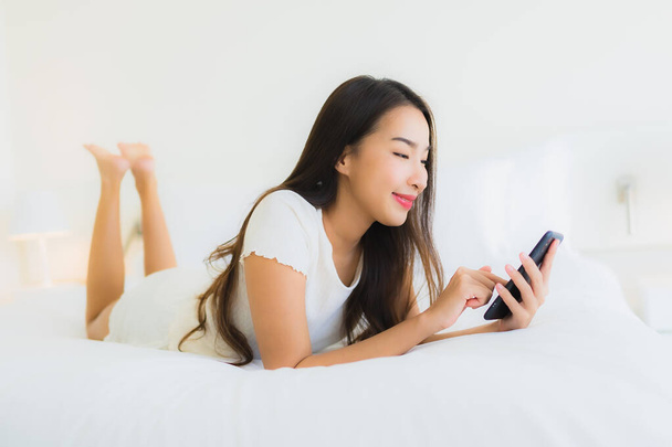 Портрет красивая молодая азиатская женщина использовать смартфон на кровати с белой подушкой одеяло в интерьере спальни - Фото, изображение