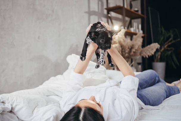 ευτυχισμένη μελαχρινή γυναίκα με κρατά ένα γαλλικό κουτάβι μπουλντόγκ στα χέρια του σε ένα κρεβάτι. - Φωτογραφία, εικόνα