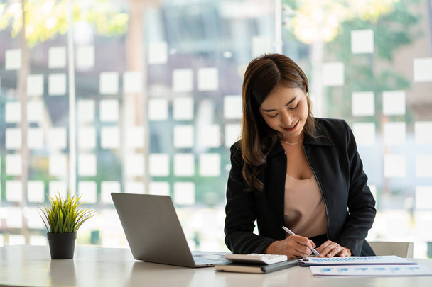 Azjatycka bizneswoman pracująca w księgowości finansowej analizuje wykres używając kalkulatora laptopa z dokumentami i trzymając długopis siedzący na krześle w biurze. - Zdjęcie, obraz