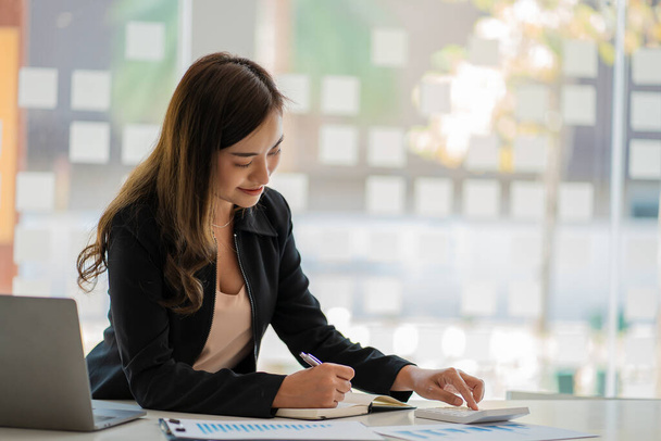 Азиатская бизнесвумен, работающая в сфере финансового учета анализирует диаграмму с помощью ноутбука-калькулятора с документами и ручкой, сидящей на стуле в офисном столе. - Фото, изображение