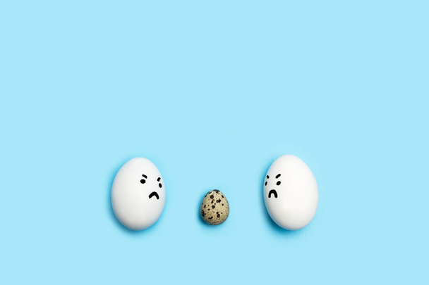 Два яйца с сердитым лицом, смотрящие на перепелиное яйцо на синем фоне - Фото, изображение