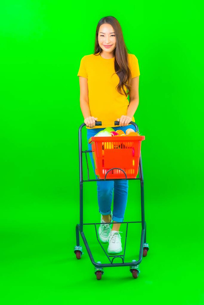 Portret mooie jonge aziatische vrouw met boodschappenmandje en kar van supermarkt op groene achtergrond - Foto, afbeelding