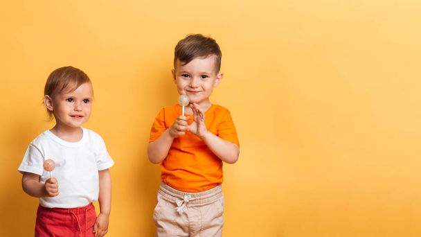 Little brother and sister are eating a sweet lollipop on a stick. Yellow background. The concept of prevention of caries and diabetes in children. Place for your text - Valokuva, kuva