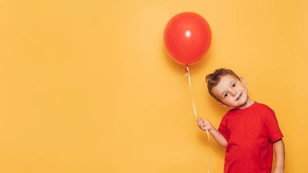 A happy Caucasian boy isolated on a bright yellow background holds a red balloon in his hands. A place for your text or advertisement. - 写真・画像