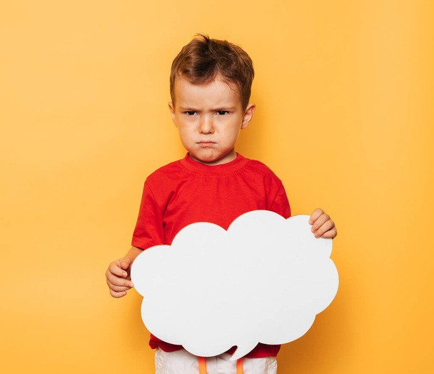 Studio portrait of a disappointed boy with a clean white board in the shape of a cloud on a bright yellow background, with a place for your text or advertising - 写真・画像