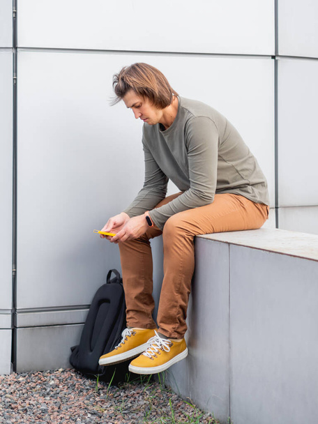 Thoughtful young man sits with smartphone on outdoor part of wall. Student with rucksack in casual clothes. Handsome man with long hair. Modern urban lifestyle. - Fotó, kép