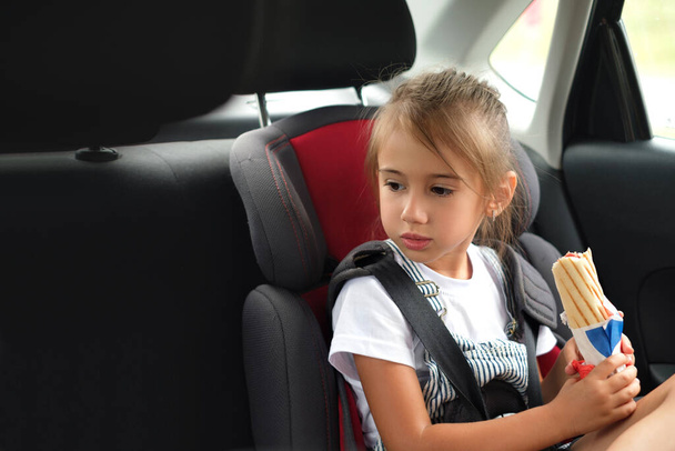 A child in a child's car seat in the back seat, fastened with seat belts, holds a bun with a sausage in his hands. A snack during the trip. The harm of fast food. A long, exhausting road journey - Photo, Image
