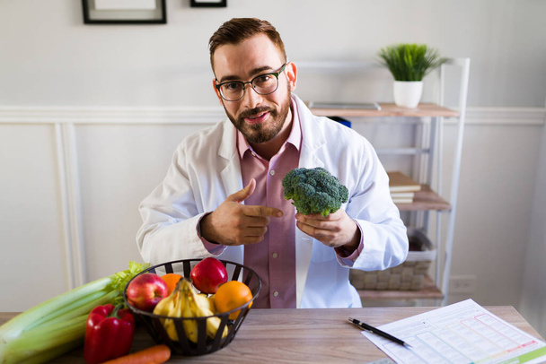 Handsome caucasian nutritionist holding a broccoli and green vegetables for a patient at his office  - Photo, Image