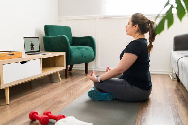Een kalme jonge vrouw die thuis yoga doet. Ontspannen vrouw doet ademhalingsoefeningen terwijl ze mediteert met een virtuele coach - Foto, afbeelding