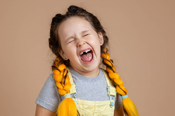 Crying little girl with yellow kanekalon pigtails, with closed eyes and opened mouth with missing tooth wearing yellow jumpsuit and gray t-shirt on beige background - Foto, imagen