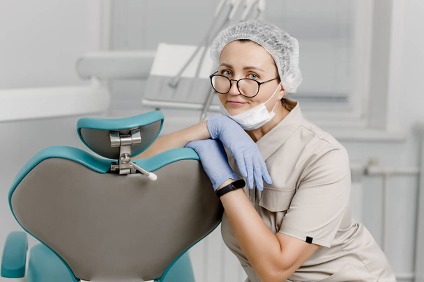 Γυναίκα οδοντίατρος με ιατρική μάσκα και καπέλο, κάθεται σε μια καρέκλα σε μια οδοντιατρική κλινική ανάμεσα σε σύγχρονο οδοντιατρικό εξοπλισμό. Πορτραίτο γυναίκας γιατρού - επαγγελματία οδοντίατρου, ορθοδοντικού - Φωτογραφία, εικόνα