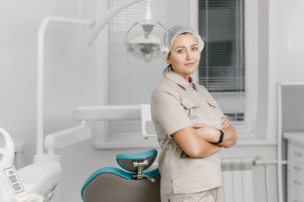 Krásná dospělá žena zubní lékař-pediatr stojí s rukama zkříženýma na hrudi v zubní klinice na pozadí moderního vybavení pro bezbolestnou zubní péči - Fotografie, Obrázek