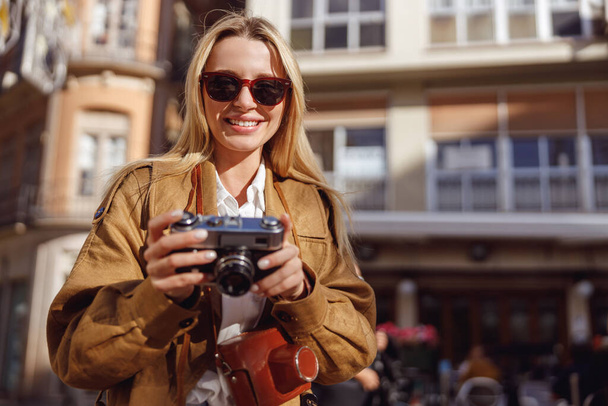 Charmante Dame steht auf der Straße in der Nähe eines Cafés und hält eine Retro-Kamera in der Hand - Foto, Bild