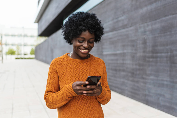 Genç, gülümseyen afro kadın sokakta yürürken akıllı telefonunu kullanıyor.. - Fotoğraf, Görsel