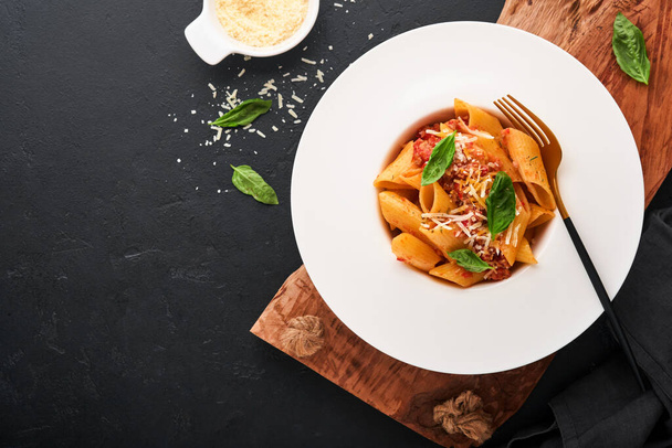 Bolognese Penne Pasta. Tasty appetizing classic italian penne pasta with parmesan, basil and Bolognese sauce in white plate on plate on black dark background.Traditional Italian cuisine. Top view. - Photo, image