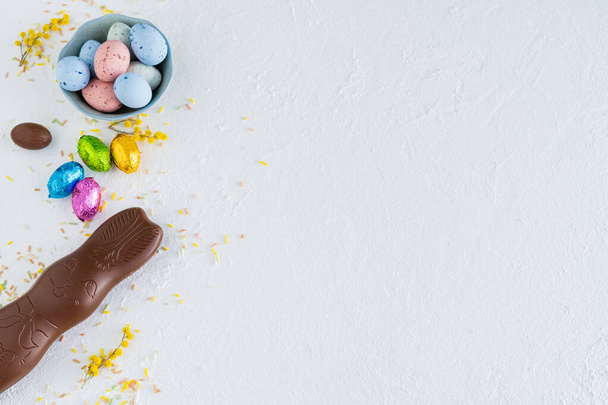 Easter eggs and chocolate bunny on white background with yellow flower decorations. Top view. Copy space. - 写真・画像