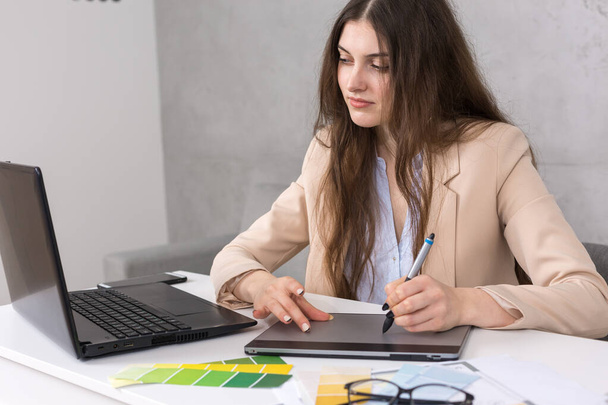 Ein junges Designermädchen, das an einem Tisch sitzt, zieht an einem Tablet. Erstellt Geschäftsberechnungen auf dem Computer. - Foto, Bild