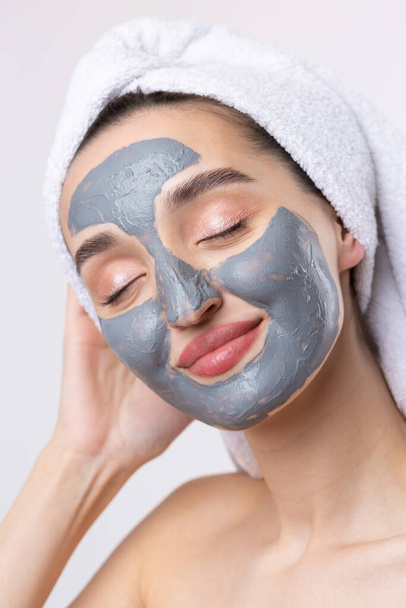 A woman with a gray clay cosmetic mask in a white towel on her head on a white background. - Foto, Imagen