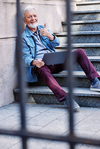 Mutlu bir son sınıf öğrencisi merdivenlerde oturuyor, dizüstü bilgisayarını tutuyor ve baş parmağını kaldırıyor.. - Fotoğraf, Görsel