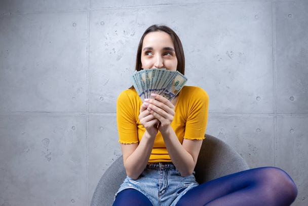 Photo of a wealthy woman in simple clothes holding a fan of dollar money isolated against a concrete wall background - Photo, image