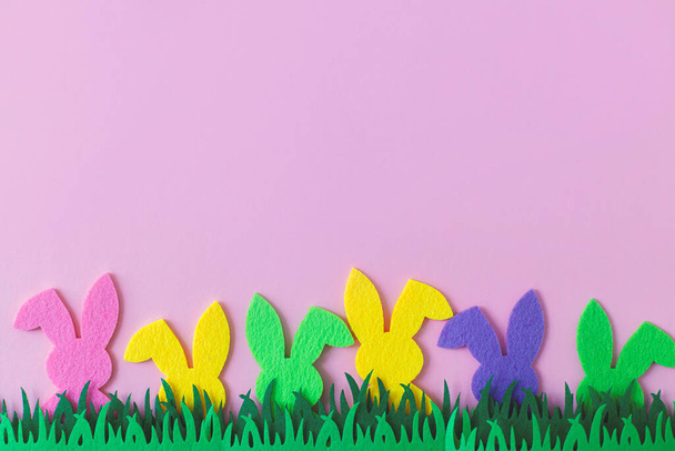 Coniglietti pasquali colorati in erba su sfondo rosa, vista dall'alto con spazio per il testo. Buona Pasqua! Rosa, giallo, verde decorazione coniglietti artificiali con erba verde. Concetto caccia pasquale - Foto, immagini