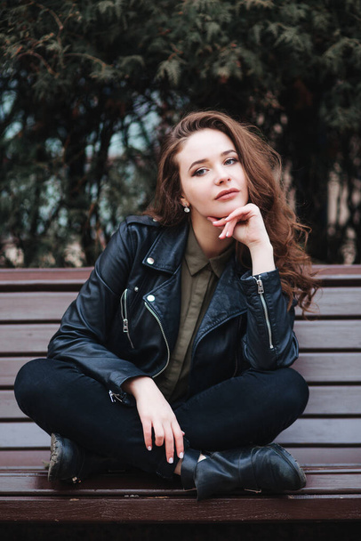 young curly stylish woman wearing black jacket sitting on a bench in city outdoor - Fotoğraf, Görsel