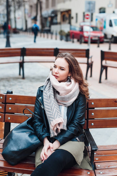 young tylish woman wearing black jacket sitting on a bench in city - Фото, зображення