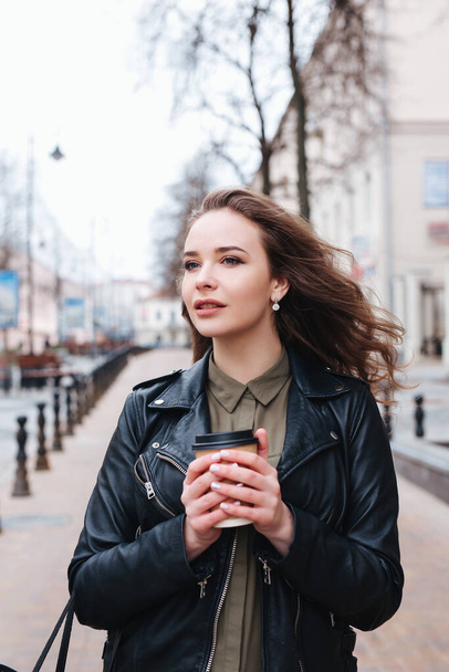 黒い革のジャケットを着た若い巻き毛の女性が歩きコーヒーを飲み街を眺め - 写真・画像