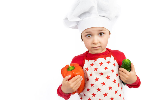 Šťastný teen chlapec v čepici šéfkuchaře drží pepř a zelené okurky, izolované na bílém pozadí. - Fotografie, Obrázek