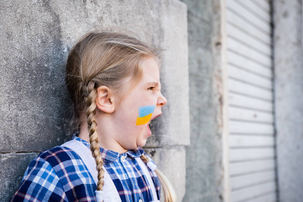Porträt eines kleinen Mädchens, das schreit und eine Fahne auf die Wange gemalt hat, die in den gelb-blauen Farben der ukrainischen Flagge gehalten ist. Konzept des Friedens und des Kinderschutzes - Foto, Bild