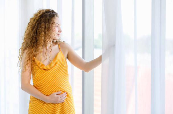 Göndör frizura boldog egészséges kaukázusi terhesség anya modell narancssárga csík anyaság hosszú ruha ruha áll mosolyogva megható nagy hasnyílás függöny látszó kívül üveg ablak hálószoba. - Fotó, kép