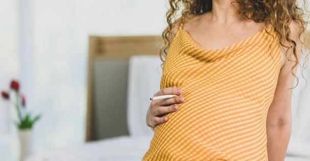 Curly hairstyle young unhappy unhealthy bad behavior Caucasian pregnancy mother in maternity long dress cloth sit on bed in bedroom holding smoking cigarette taking risk and danger to unborn child. - Foto, Imagen