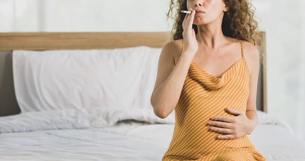Curly hairstyle young unhappy unhealthy bad behavior Caucasian pregnancy mother in maternity long dress cloth sit on bed in bedroom holding smoking cigarette taking risk and danger to unborn child. - Photo, Image