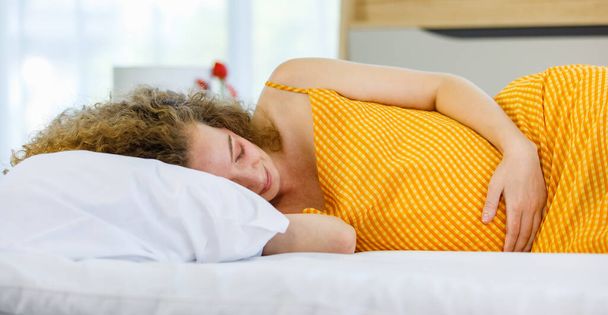 Curly hairstyle young tired exhausted sleepy Caucasian pregnancy mother in orange maternity long dress cloth lay down holding hand on big belly tummy with love sleeping on pillow and bed in bedroom. - Foto, Imagem